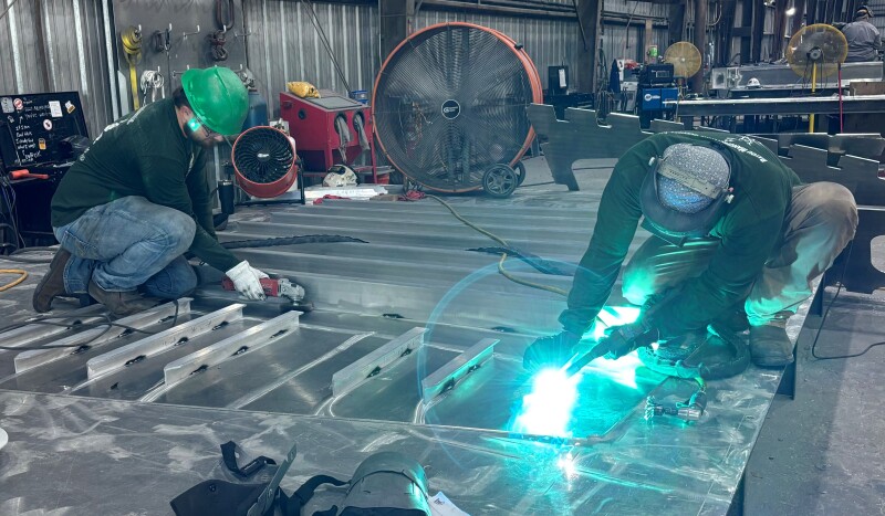 Two metal fabrication workers in a workshop weld and grind a large aluminum structure on the floor while wearing protective helmets, gloves, and safety gear, with industrial equipment and ventilation fans visible in the background.